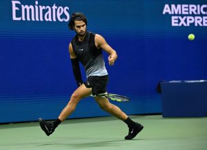 A tennis player in a black sleeveless top and black shorts runs to hit a forehand on a blue court, with an Emirates banner and a green ball in view.