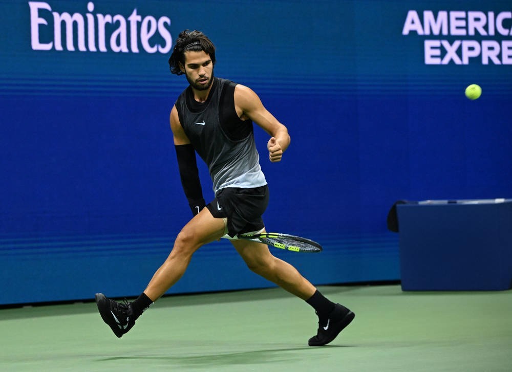 A tennis player in a black sleeveless top and black shorts runs to hit a forehand on a blue court, with an Emirates banner and a green ball in view.