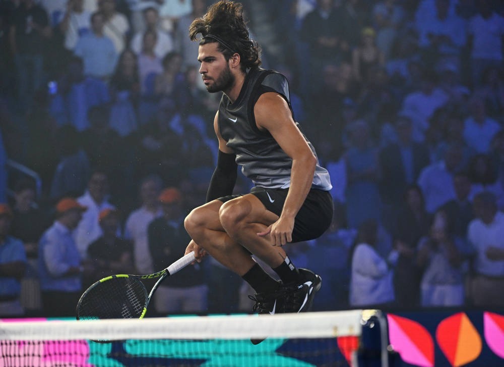 Tennis player in a black sleeveless top and shorts crouching mid-rally with a racket, on court at a night match before a blurred crowd, net in foreground.