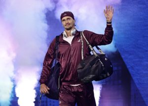 A tennis player in a maroon Adidas tracksuit with a headband and headphones waves to the crowd amid purple stage smoke.
