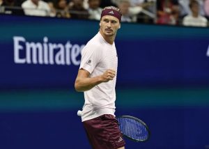 Tennis player in maroon headband and shorts, clenching fist and holding racquet on court at the US Open, Emirates sign behind him