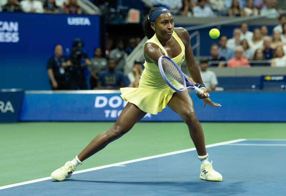 Tennis player in yellow dress and blue headband swinging a racket on the court at the US Open, with a ball in the air.