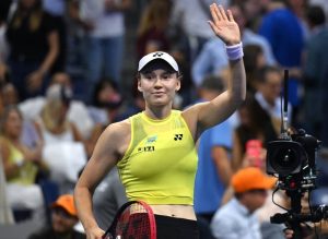 Tennis player in yellow Yonex top and white visor waving to the crowd at the 2026 US Open, holding a red racket, with a TV camera nearby