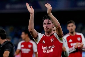 Footballer in red and white Arsenal kit applauding fans at night, with teammates behind him, Emirates sponsor visible
