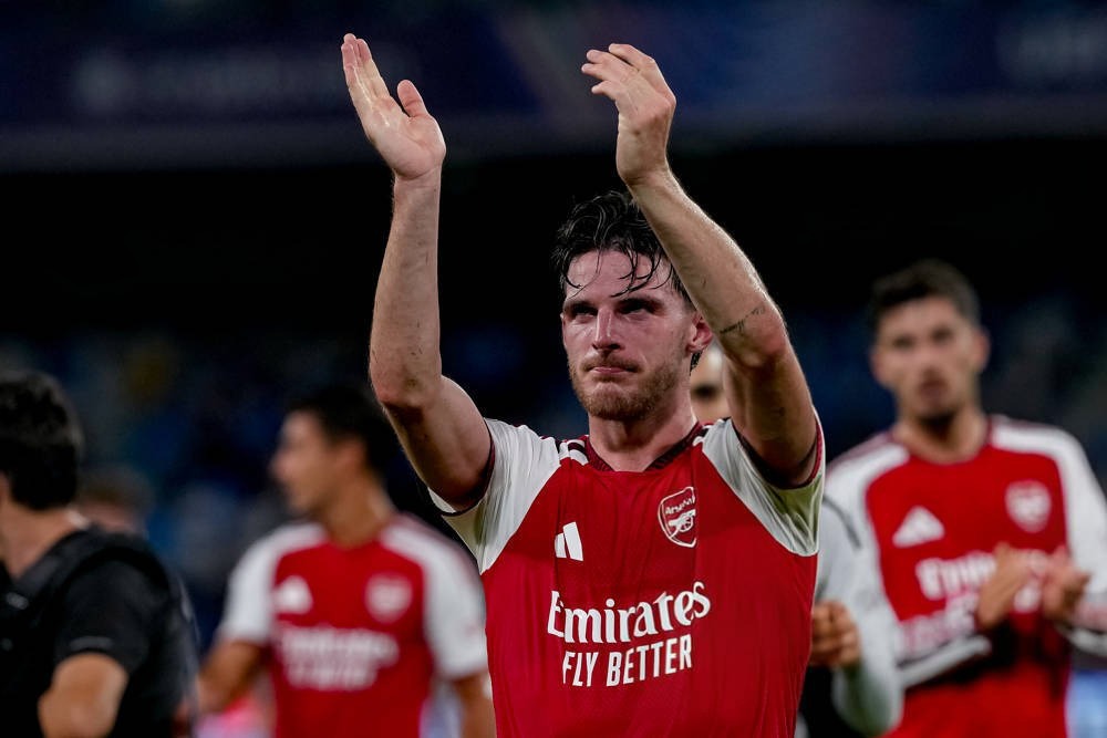 Footballer in red and white Arsenal kit applauding fans at night, with teammates behind him, Emirates sponsor visible
