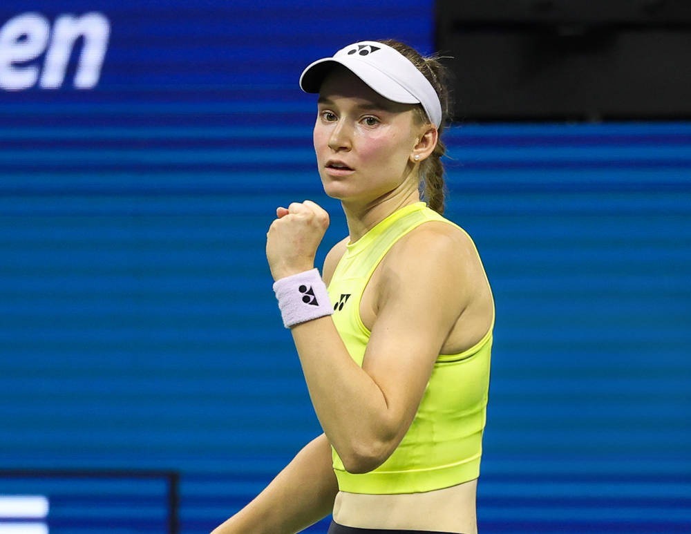 Tennis player in a yellow top and white visor with Yonex logo, clenching fist on court at the US Open, celebrating a point.