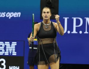 Tennis player in black mesh dress and silver chain necklace clenching her fist and holding a green racquet at the US Open court
