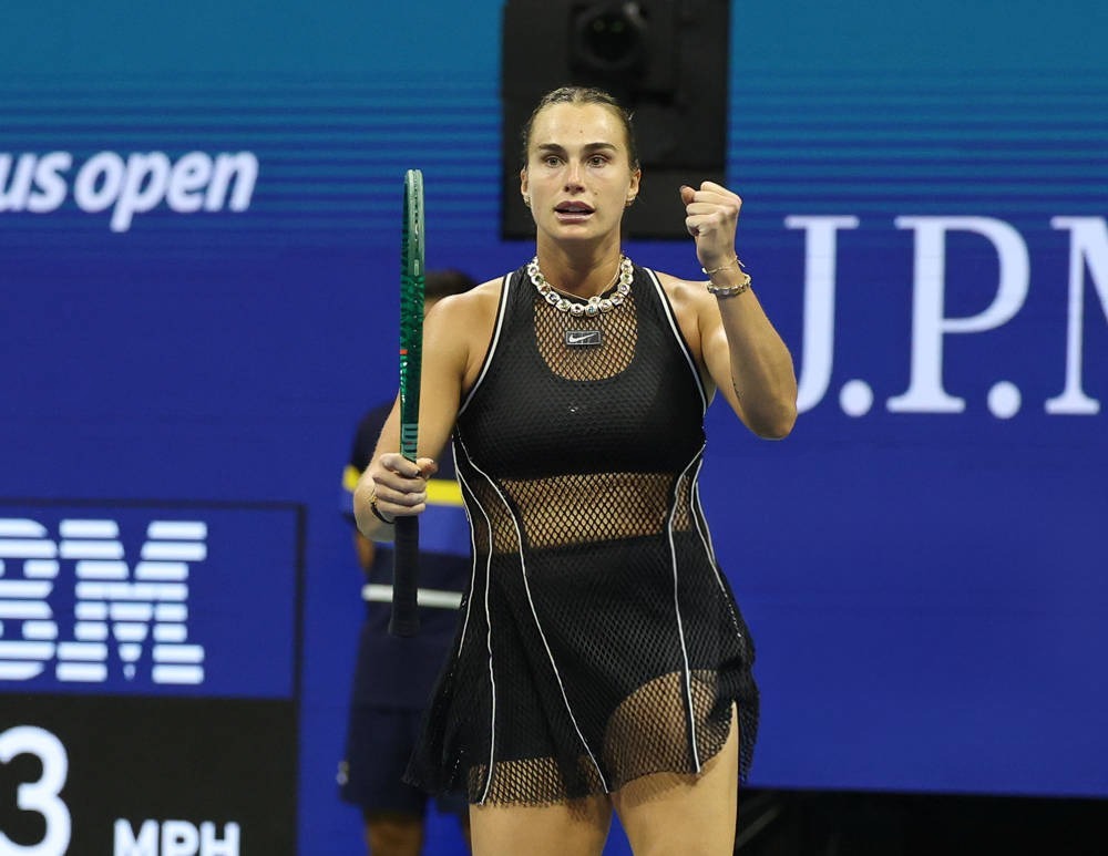 Tennis player in black mesh dress and silver chain necklace clenching her fist and holding a green racquet at the US Open court