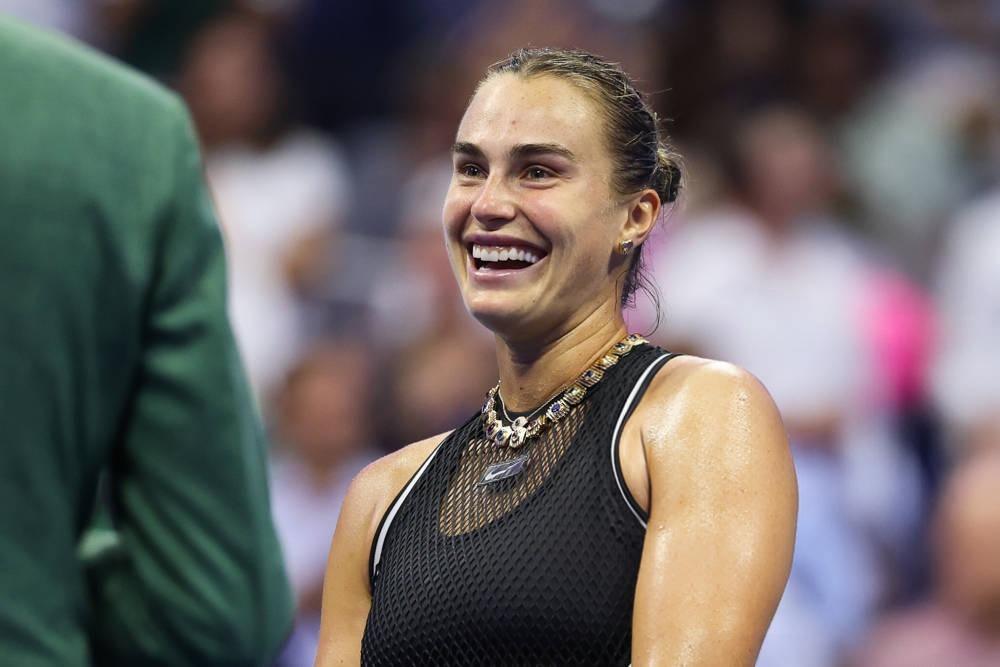 Tennis player with slicked-back hair and a black mesh top, smiling broadly and wearing a chunky gold necklace, at a US Open match setting
