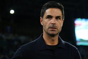 A man with short dark hair and stubble in a dark polo shirt, looking to the side at a stadium with blurred lights and a screen behind him