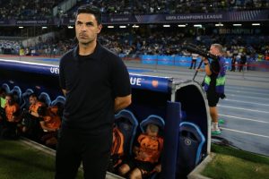 Man in black polo standing by the dugout at a UEFA Champions League match, Arsenal substitutes seated behind him in orange and black.