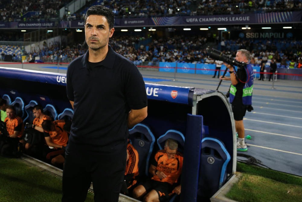 Man in black polo standing by the dugout at a UEFA Champions League match, Arsenal substitutes seated behind him in orange and black.