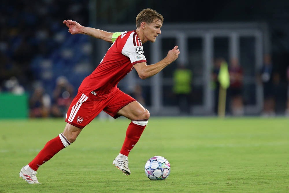 Martin Odegaard in red Arsenal kit and captain's armband, running with the ball during a Champions League match.