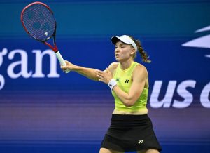 Tennis player in yellow Yonex top and black skirt swinging a red Yonex racket on a blue US Open court