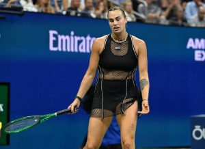 Tennis player in a black mesh outfit holding a green racquet, grimacing on court at the US Open with Emirates and American Express signage behind her.