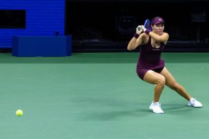 Tennis player in a maroon dress and cap hitting a backhand on a green court, with a yellow ball in flight at the US Open