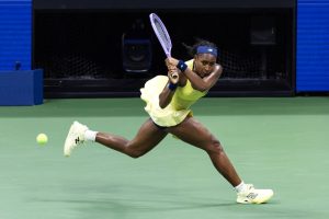 Tennis player in a yellow dress and blue headband hitting a backhand on a green court, with a New Balance racket and shoes