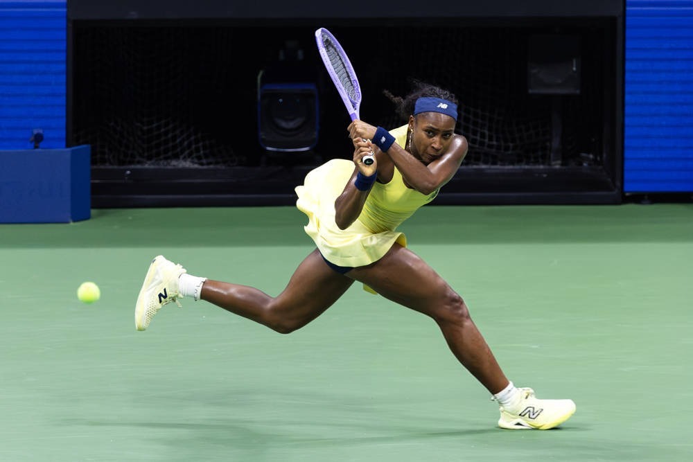 Tennis player in a yellow dress and blue headband hitting a backhand on a green court, with a New Balance racket and shoes