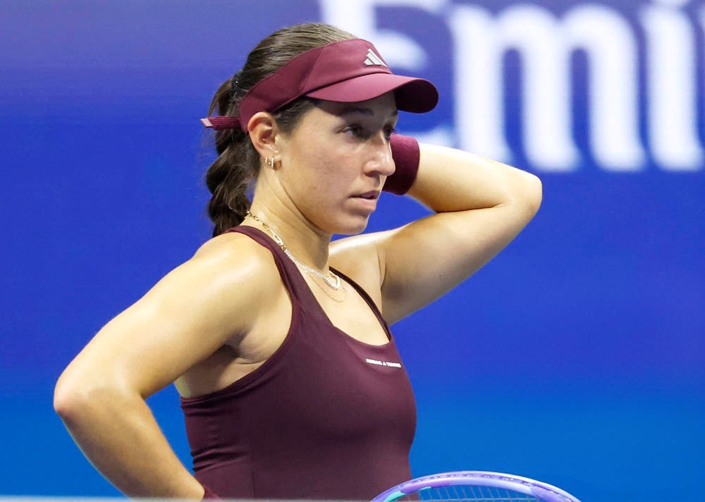 Tennis player in a maroon visor, tank top and wristband, hand on head during a match with a blue court backdrop and a racket visible