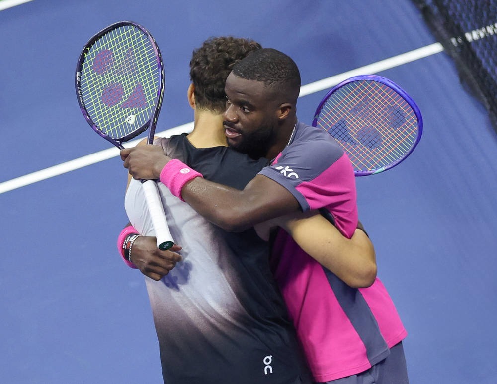 Tennis players embracing at the net after a match, one in a pink-and-grey shirt with a Yonex racket, the other in black with an On logo.