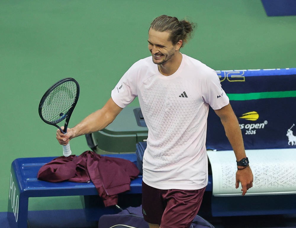 Tennis player in a pink Adidas shirt and maroon shorts, smiling and holding a racquet courtside at the 2026 US Open.