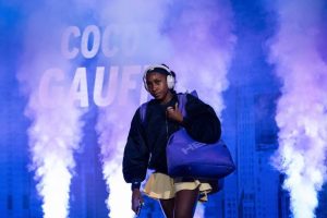Tennis player in a dark jacket and yellow skirt, headphones on, carrying a purple bag, walking through purple smoke with "COCO GAUFF" on the backdrop