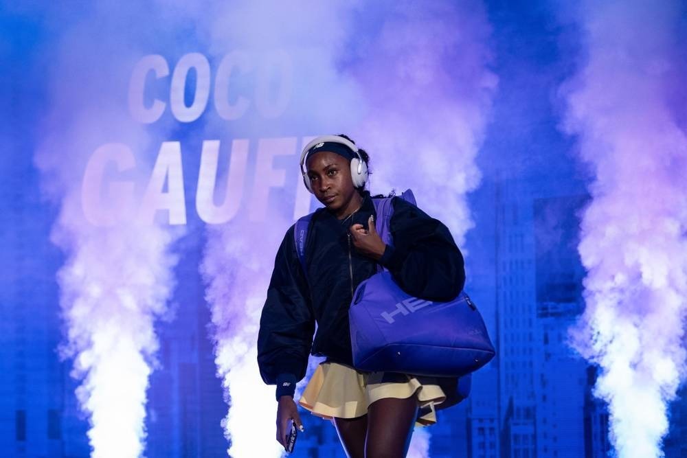 Tennis player in a dark jacket and yellow skirt, headphones on, carrying a purple bag, walking through purple smoke with "COCO GAUFF" on the backdrop