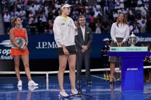 Elena Rybakina in a white jacket and visor speaks into a microphone at the US Open trophy presentation, with a runner-up beside her holding a silver plate.