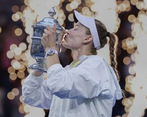 A tennis player in a white visor and jacket kissing a silver championship trophy amid glowing sparkler-style lights at a night ceremony