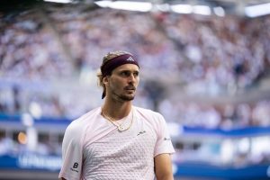 Tennis player in a pink Adidas shirt and maroon headband at a stadium, looking to the side with crowds blurred behind him.