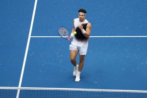 A tennis player in a white headband and sleeveless shirt, holding a blue racket and about to strike a yellow ball on a blue court