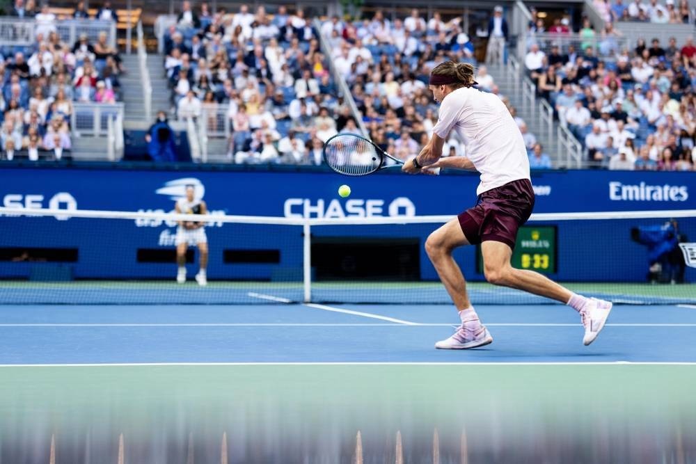 Tennis player in a white shirt and maroon shorts hitting a forehand on a blue hard court at a US Open night match, with Chase and Emirates signage.