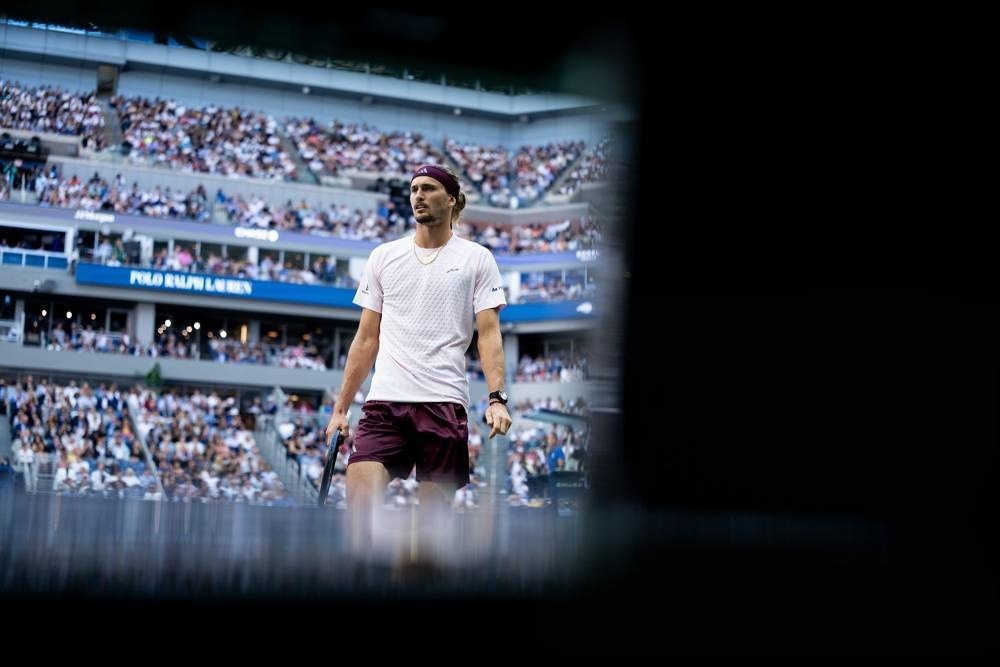 A tennis player in a white shirt and maroon shorts holds a racket on a stadium court, with crowds in the stands behind him.