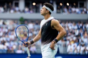 Tennis player in a black-and-white sleeveless On shirt and white headband holding a blue racket, looking up at a packed stadium crowd