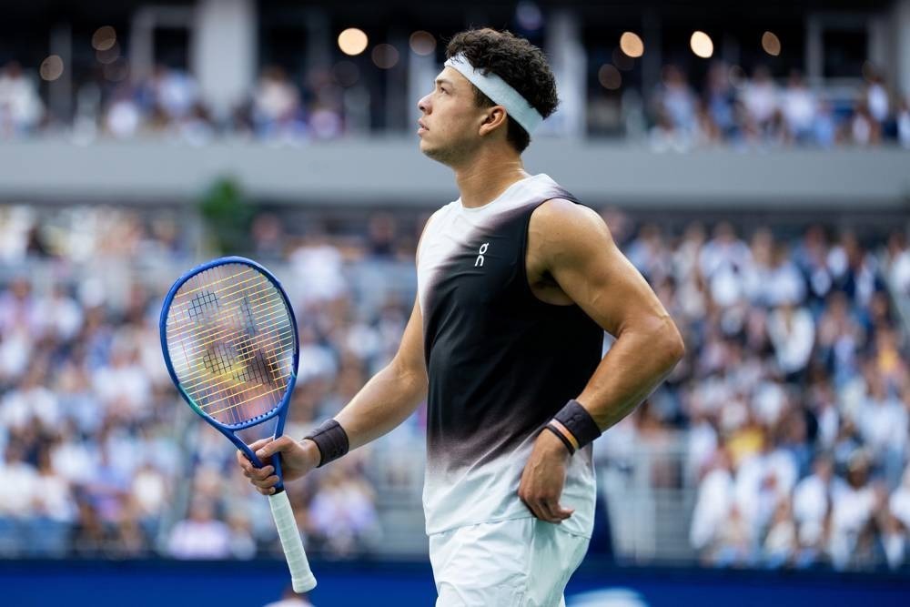 Tennis player in a black-and-white sleeveless On shirt and white headband holding a blue racket, looking up at a packed stadium crowd