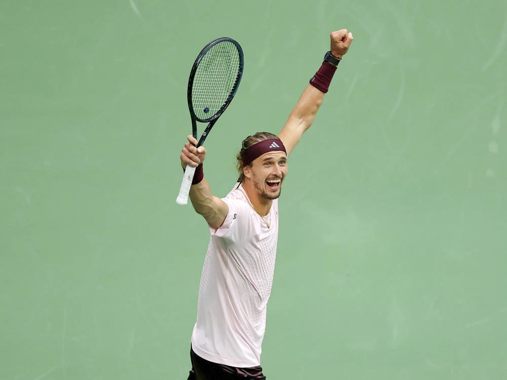 Tennis player in a white shirt and maroon headband, arms raised and fist clenched while holding a racquet on a green court, celebrating a US Open win