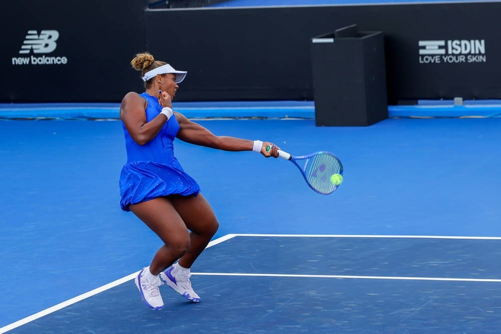 A tennis player in a blue dress and white visor hits a forehand on a blue court, with New Balance and ISDIN signage behind her