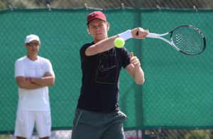 Tennis player in dark navy Nike shirt and red cap hitting a forehand on a green-fenced court, with a coach in white watching behind him.