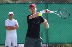 Jannik Sinner in a navy Nike shirt and maroon cap hits a forehand on a green-fenced court, with a man in white watching behind him.