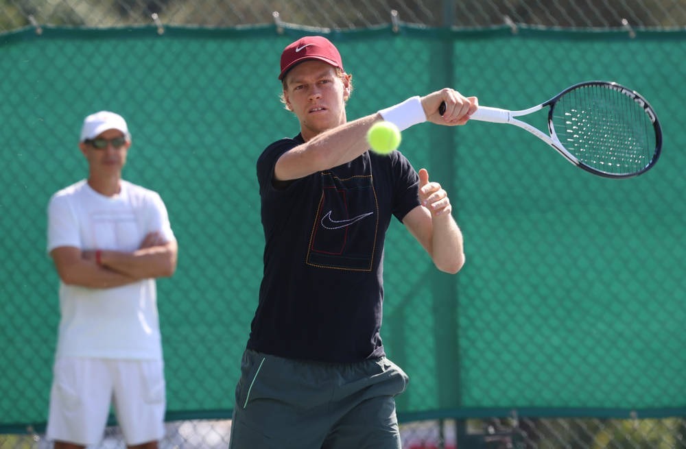 Jannik Sinner in a navy Nike shirt and maroon cap hits a forehand on a green-fenced court, with a man in white watching behind him.