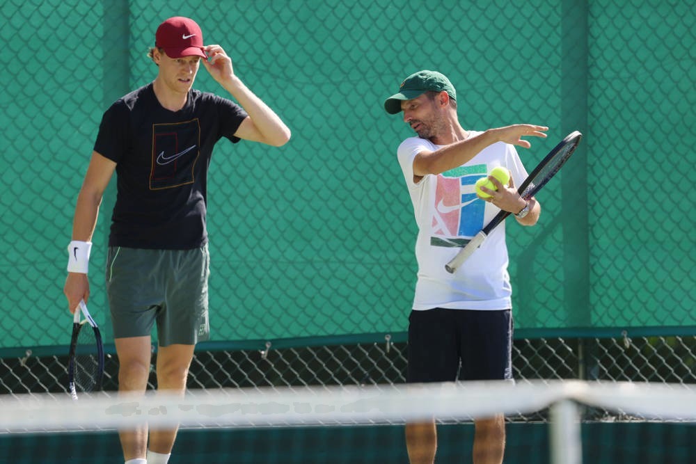 Two men on a green tennis court: a player in a red cap and black Nike shirt, and a coach in a green cap pointing with a racket and ball, during practice.