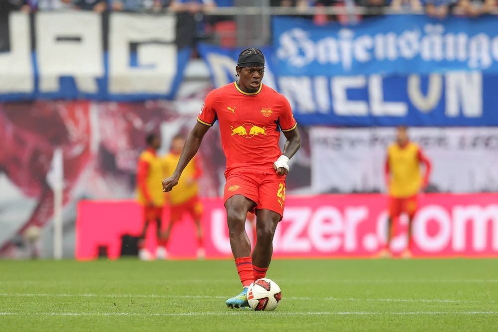 Player in red RB Leipzig kit, number 24, with black headband and wrist tape, dribbling the ball on the pitch in a stadium.