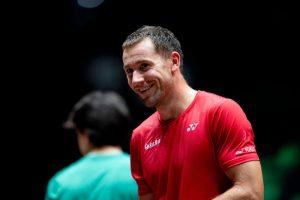 Smiling man in a red Yonex shirt at a tournament venue, with a blurred figure in green in the foreground. Likely Casper Ruud at the Shanghai Masters.
