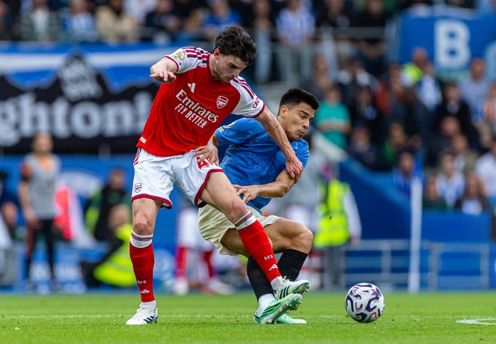 Declan Rice in red Arsenal kit with Emirates sponsor battles for the ball against a blue-shirted opponent on a sunny pitch.