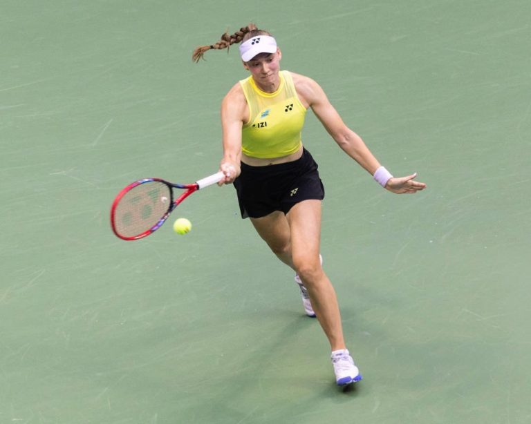 Elena Rybakina in yellow top and black skirt hitting a forehand on a green court at a tennis tournament