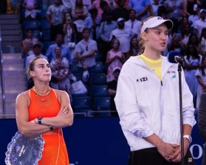 Two tennis players at a press conference: one in an orange top holding a crystal trophy, the other in a white Yonex jacket speaking into a microphone, with s…