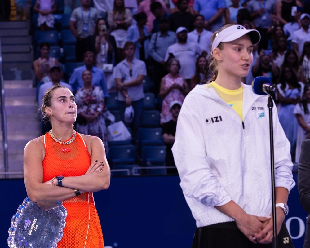 Two tennis players at a press conference: one in an orange top holding a crystal trophy, the other in a white Yonex jacket speaking into a microphone, with s…