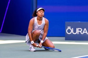 Tennis player in white visor and grey outfit crouching on court, head bowed, holding a blue racket, with an OURA sign behind her.