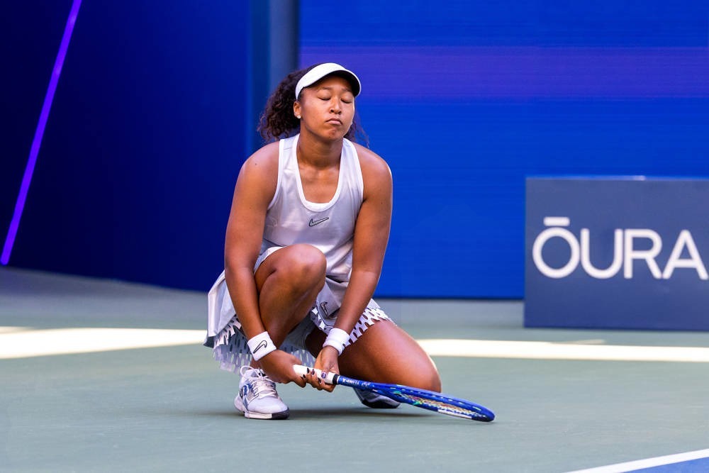 Tennis player in white visor and grey outfit crouching on court, head bowed, holding a blue racket, with an OURA sign behind her.
