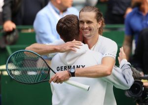 Tennis players embracing on court; one wears a white Germany jacket and holds a teal racket, the other smiles in white.